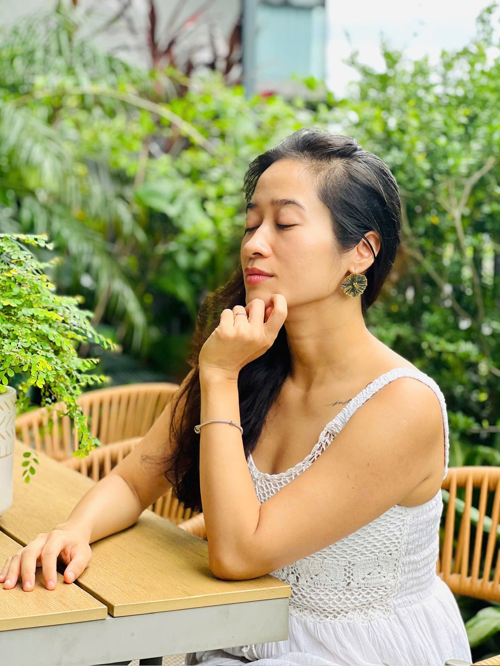 Người phụ nữ tóc đen nhắm mắt thư giãn, mặc váy ren trắng tinh khôi, ngồi bên bàn gỗ ngoài trời